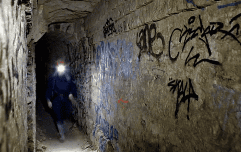 Underground explorer with headlamp walking through a graffiti-covered tunnel in the restricted areas of the Paris Catacombs